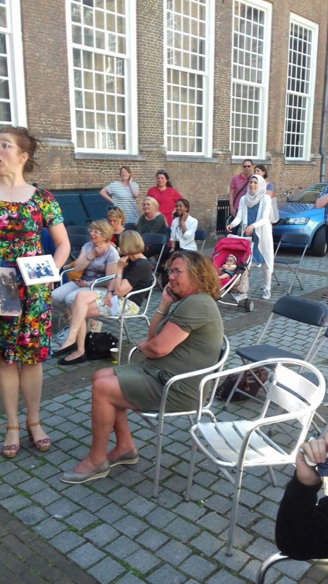 Publiek op stoelen op de binnenplaats luistert naar een vertelster met een ingelijste foto
