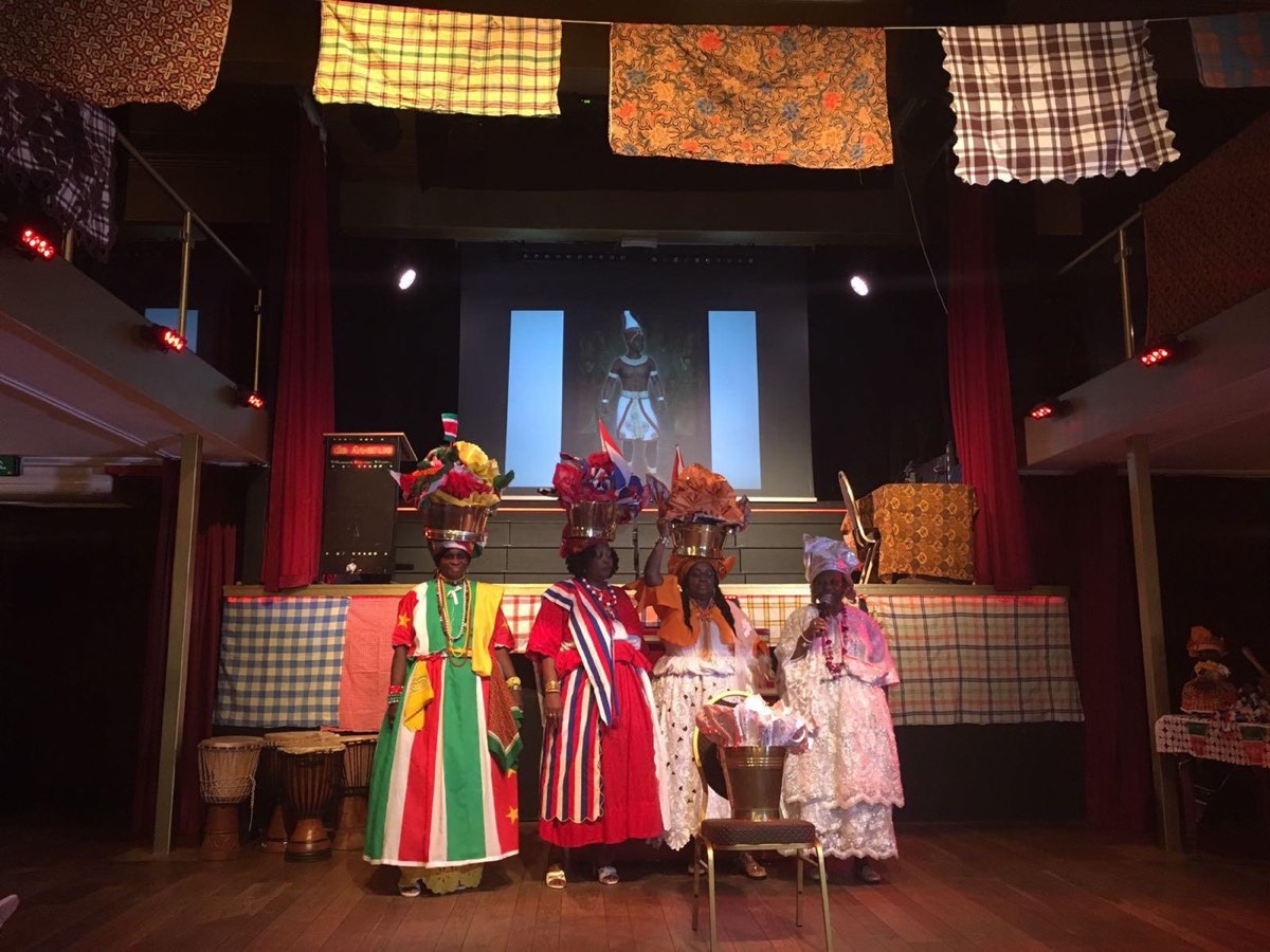 Vier vrouwen in Surinaamse koto en angisa op het podium, met versierde bekkens op het hoofd